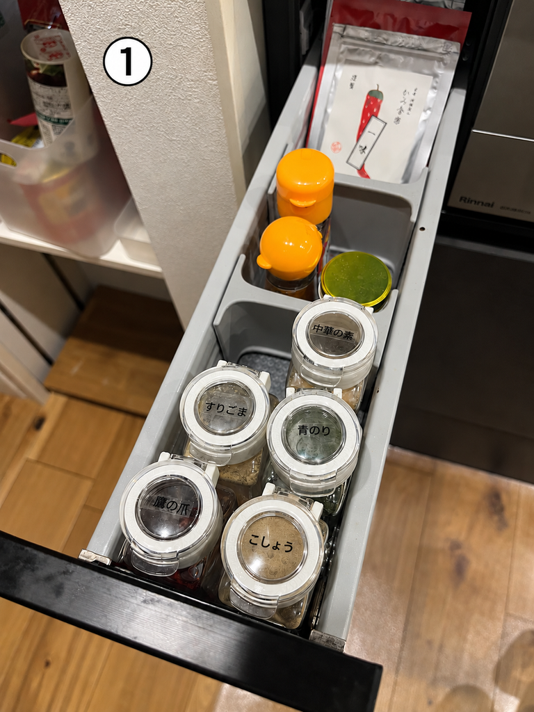 コンロ横の細い引き出しに並べた調味料。手前には「こしょう」「鷹の爪」「すりごま」「青のり」「中華の素」のラベルを付けた透明の保存容器があり、奥にはオレンジ色のふたの容器や袋入りの調味料を収納している。